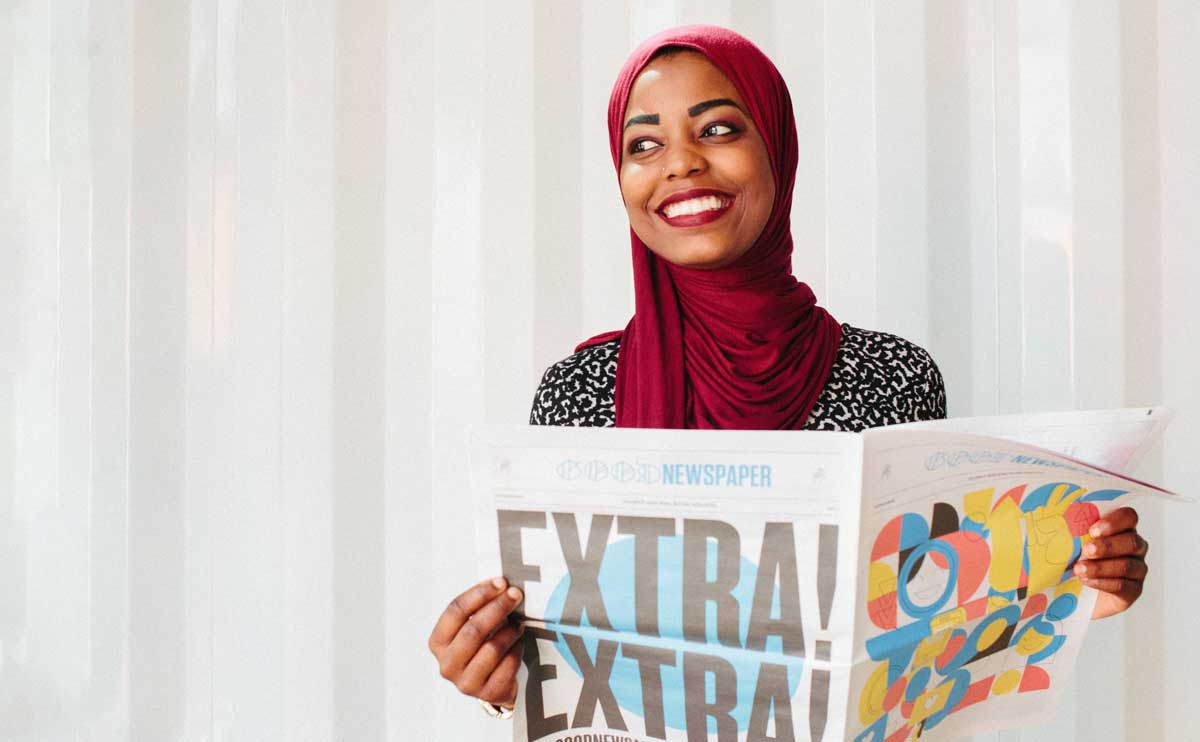 A woman in a hijab smiles as she reads a colorful happy newspaper: The Goodnewspaper