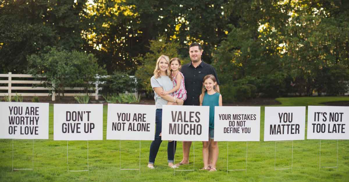 Woman stands with her family in front of several white signs that say things like "Don't Give Up", "You Are Not Alone", "You Matter", and "Your Mistakes Do Not Define You"