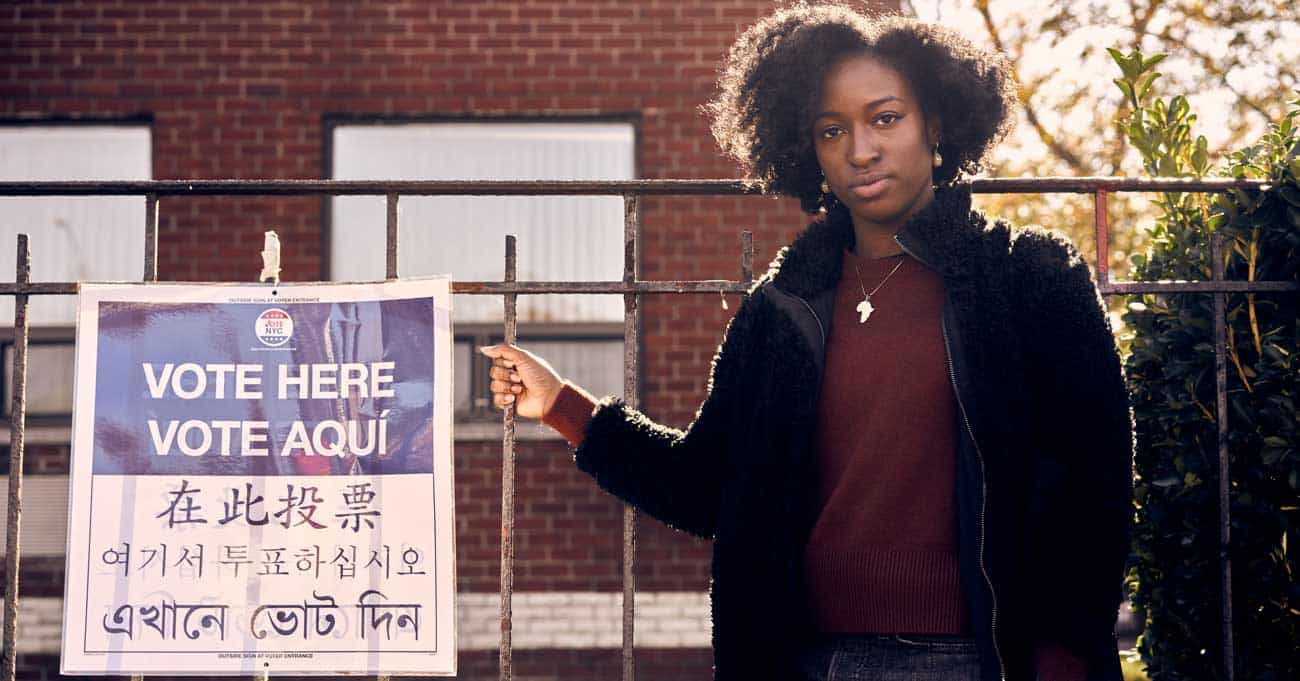 A Black woman stands in front of a sign that says VOTE HERE in multiple languages