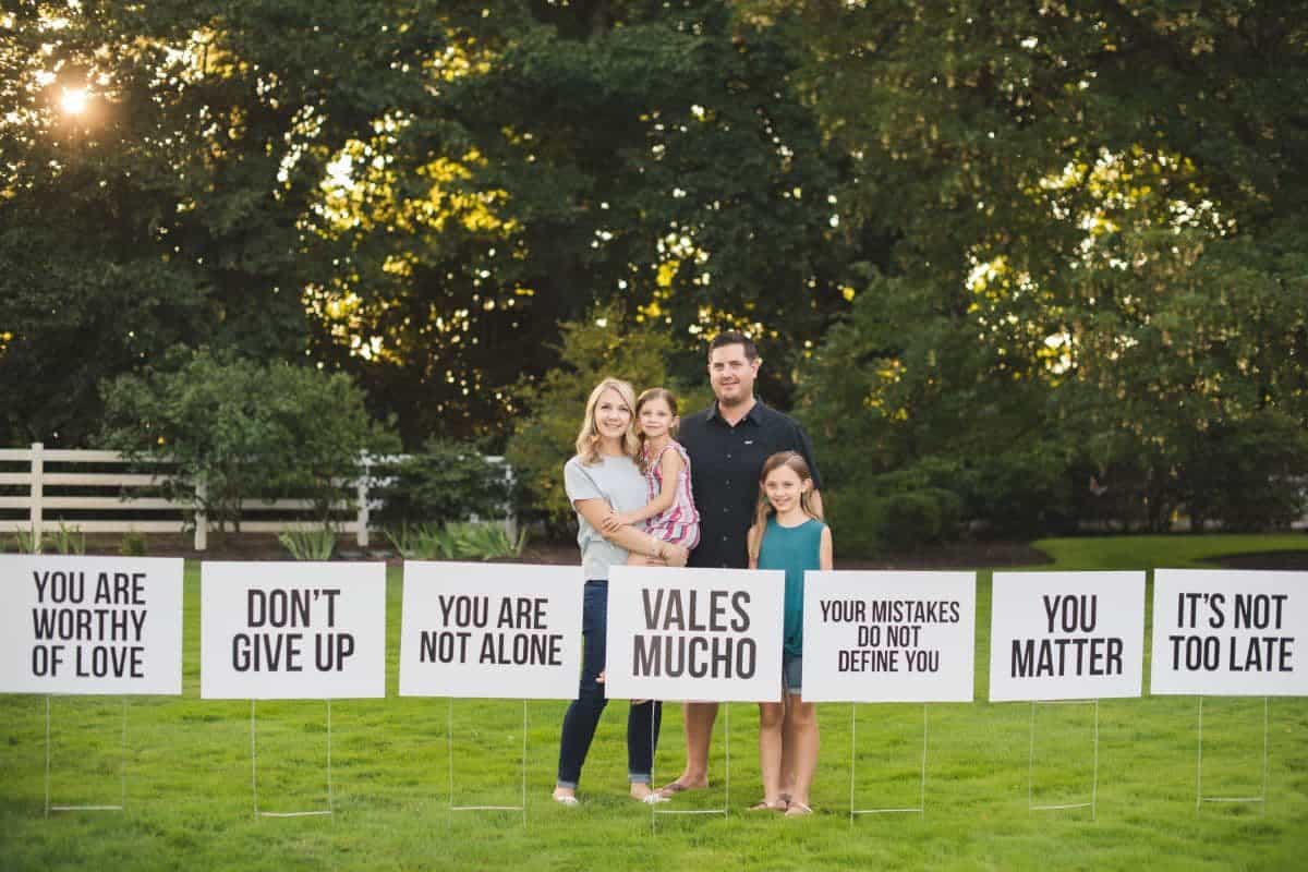 Woman stands with her family in front of several white signs that say things like Don't Give Up, You Are Worthy of Love, You Are Not Alone, Vales Mucho, Your Mistakes Do Not Define You, You Matter, It's Not Too Late