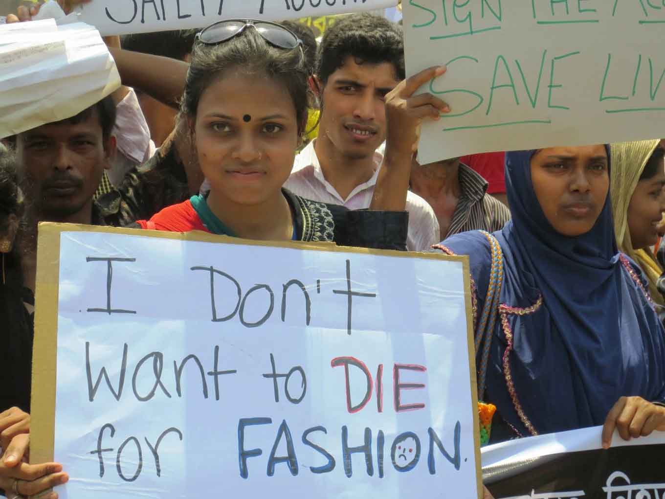 A woman in Bangladesh stands in a crowd holding a sign that says, "I don't want to DIE for FASHION"