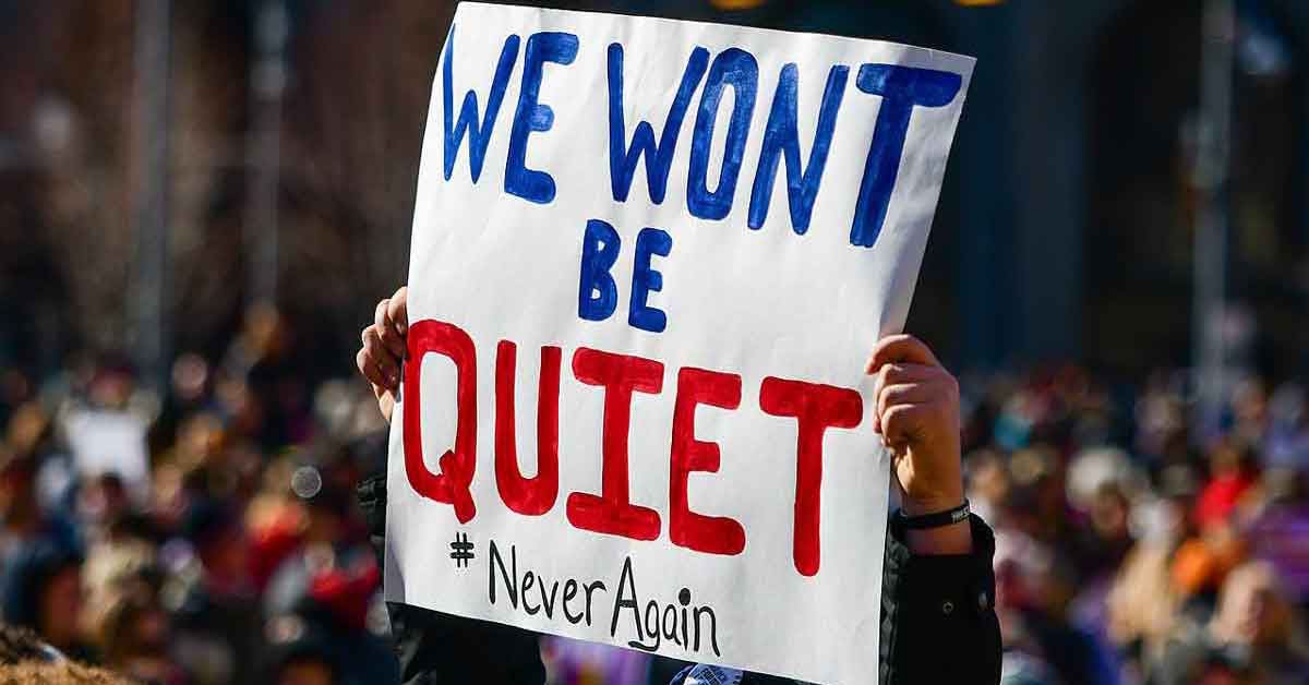 A poster stands among a crowded gun safety March for Our Lives protest, reading "We won't be quiet. #NeverAgain."