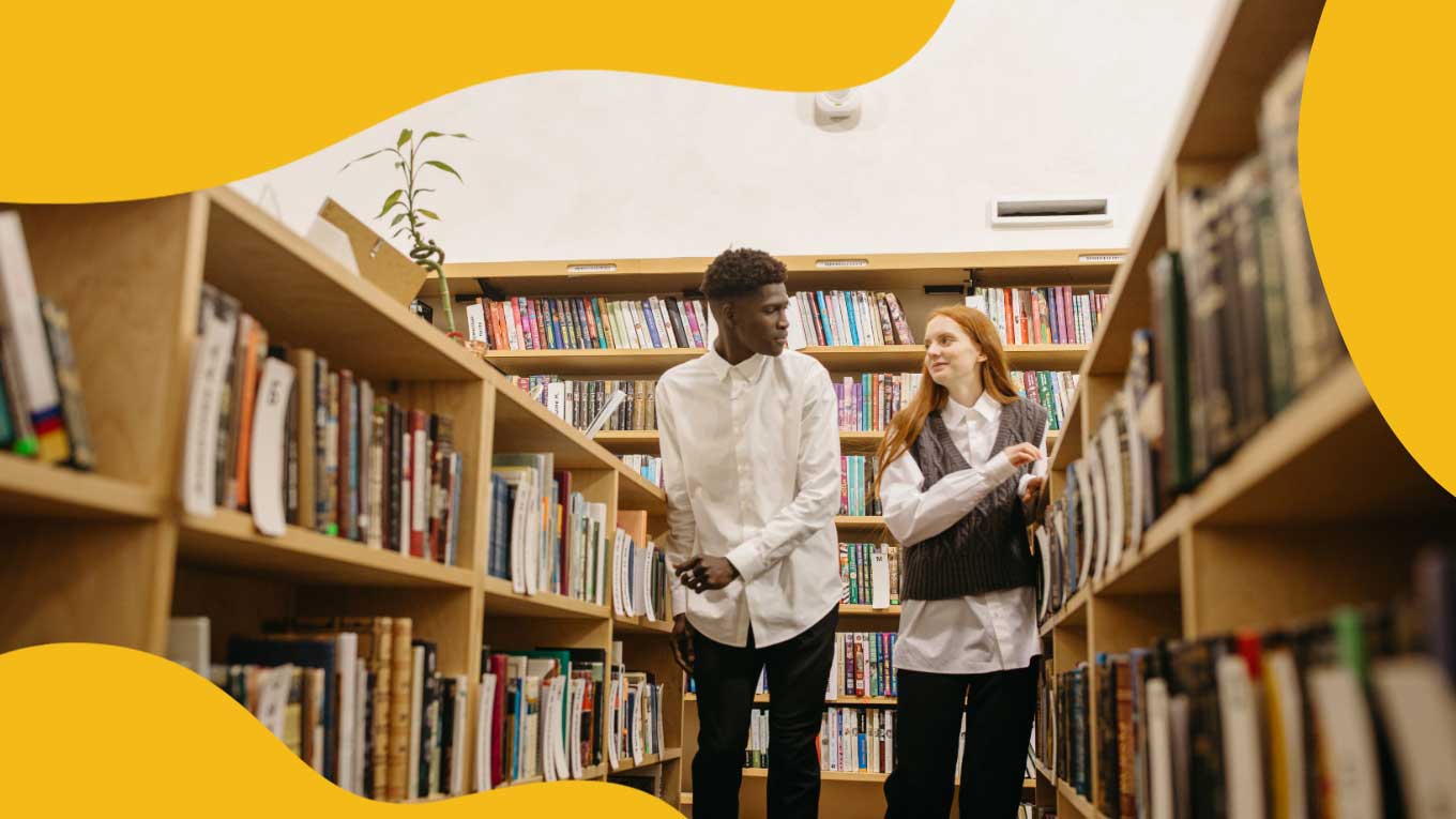 A young Black man in a white shirt and black pants talks to a young white girl with red hair and a gray sweater vest. They walk through the bookshelves in a library.