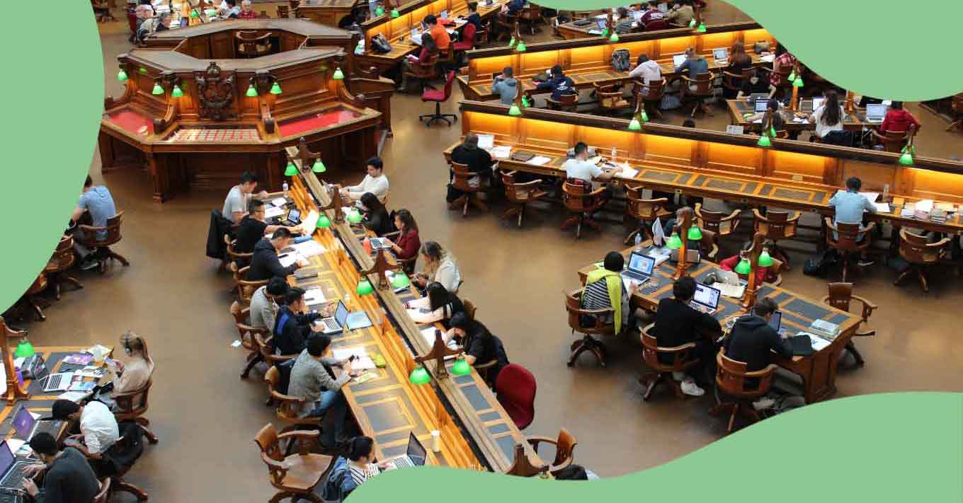 A birds-eye-view shows people working in long tables with green lamps inside a library. 