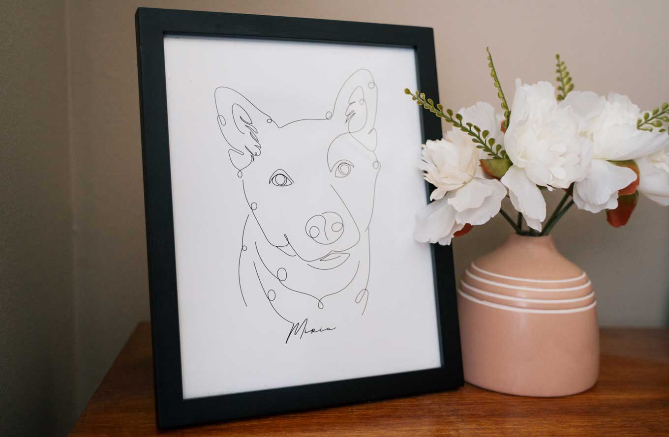 A contour-style line drawing of a dog is in a black frame. It sits next to a bouquet of white flowers.