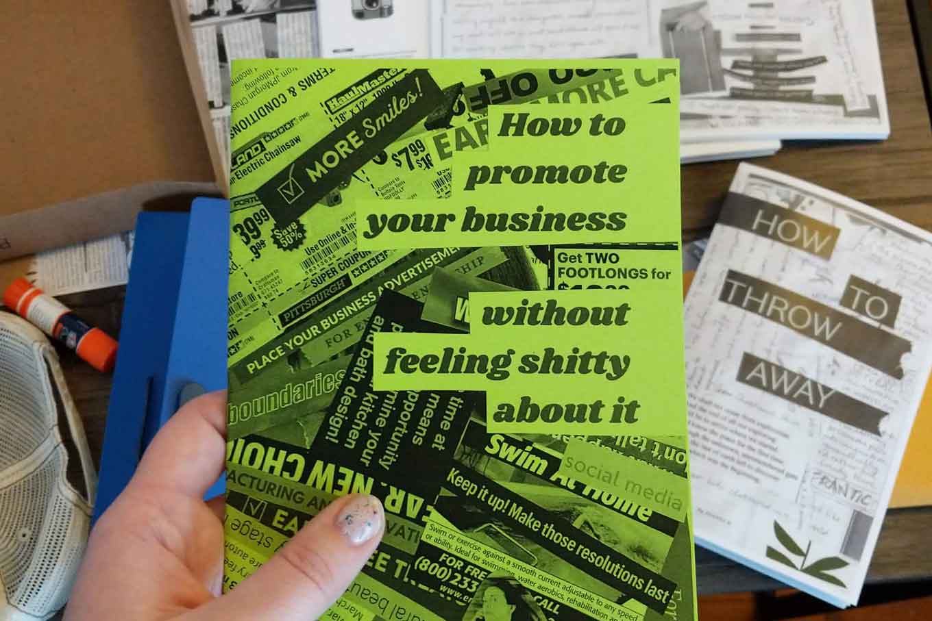 A hand holds up a copy of the zine: "How To Promote Your Business Without Feeling Shitty About It."