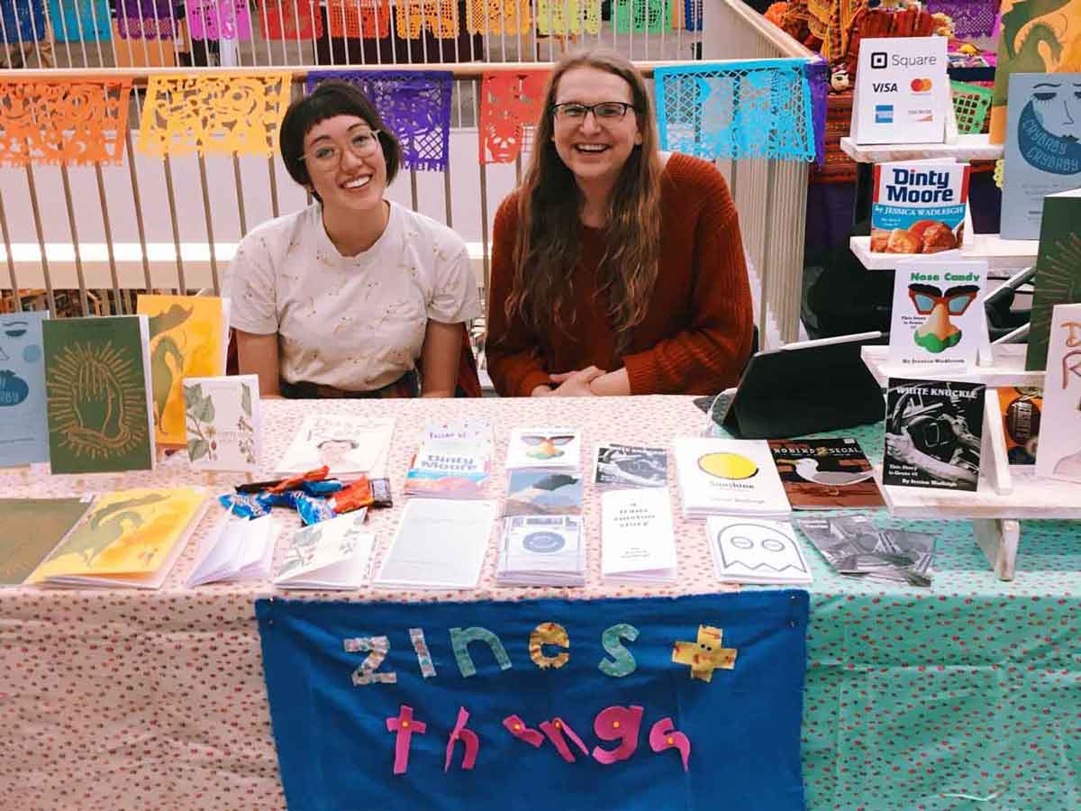 Two women sit at a colorful table, smiling. A number of zines sit atop the table.