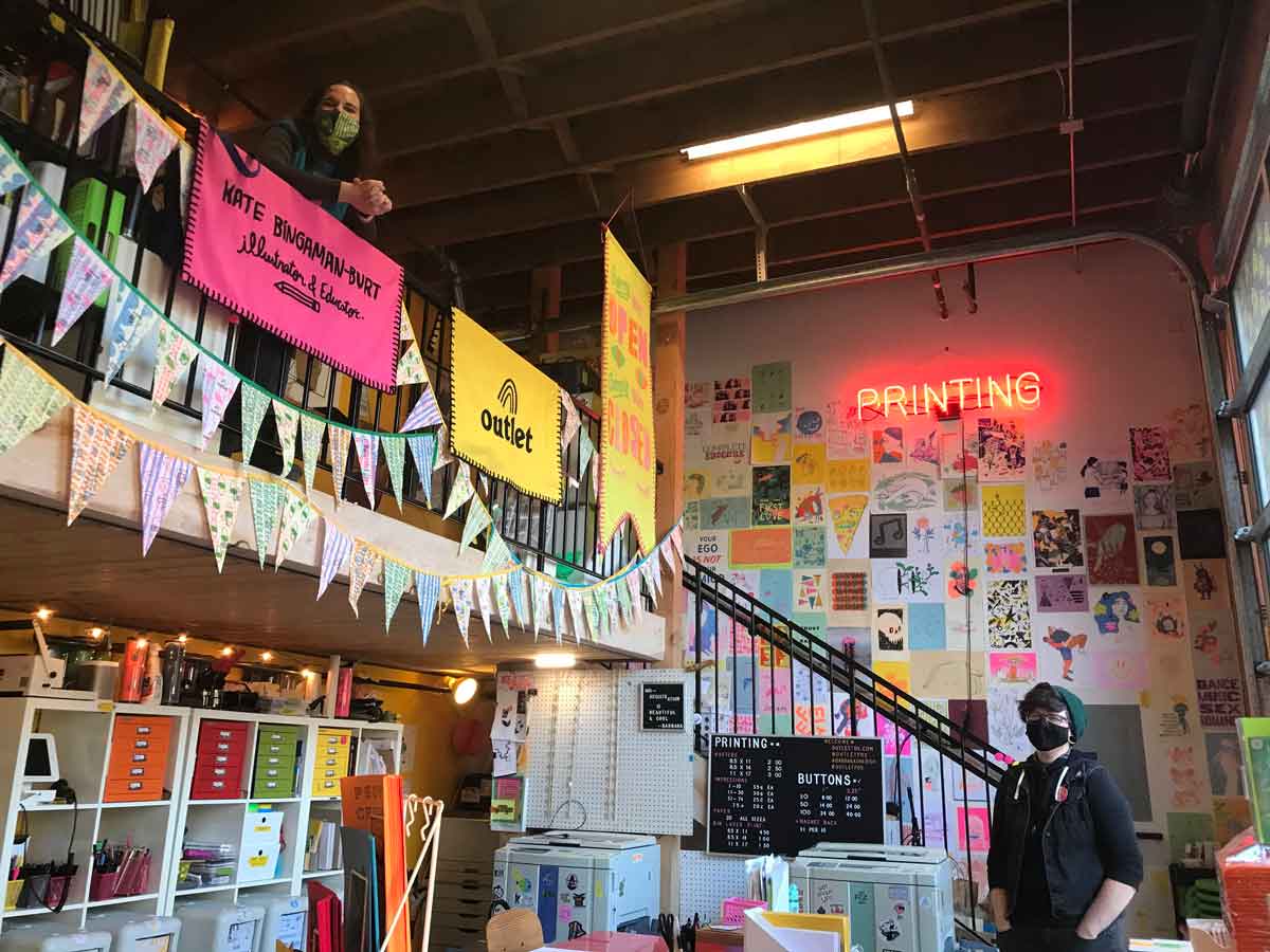 A colorful room filled with prints and zines. On the wall, a pink neon sign reads "printing." Two people stand in the room wearing masks.