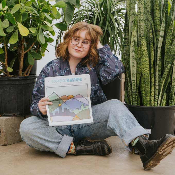 A person poses with the Goodnewspaper, surrounded by green plants