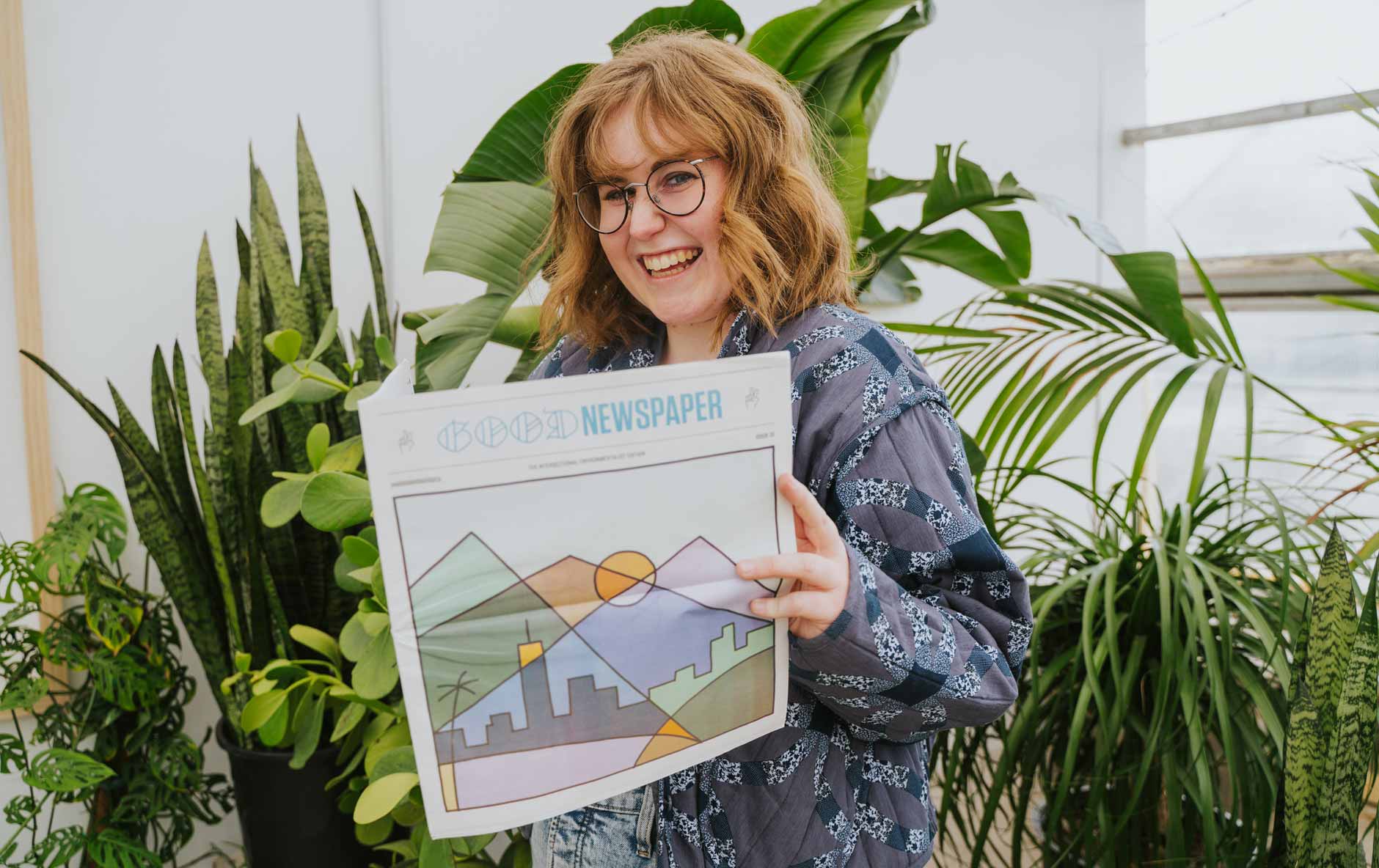 A person smiles while holding the Goodnewspaper: a colorful print newspaper filled with good news