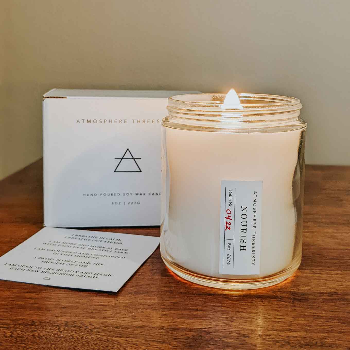 Atmosphere Threesixty glass candle that says Nourish, with its packaging behind it