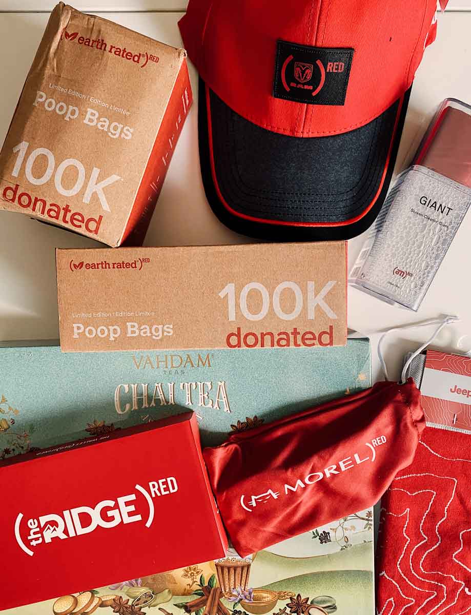 Several Product (RED) products, laid out on a table, including Earth Rated Poop Bags, a RAM hat, a pair of Morel sunglasses, Vahdam chai tea, Jeep socks, and a Ridge wallet
