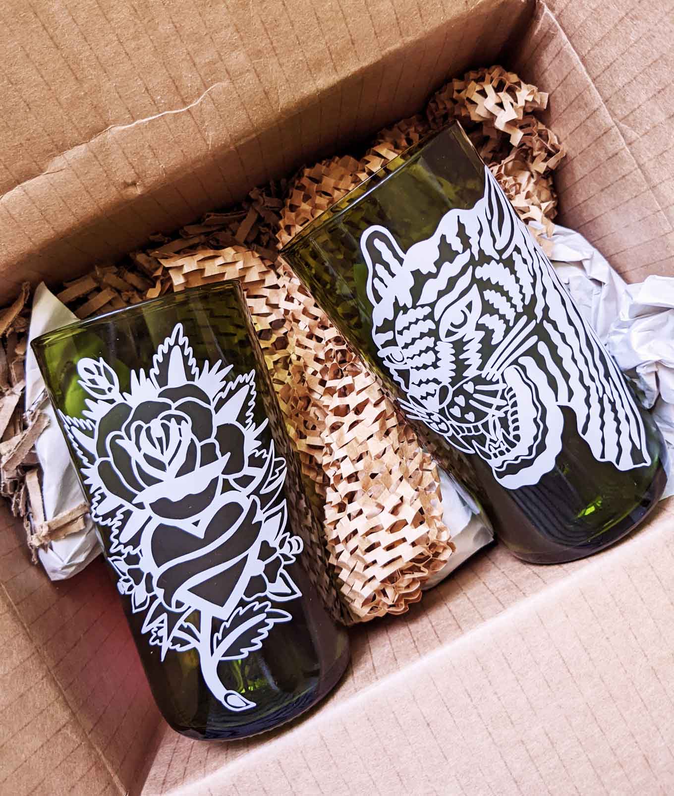 Two green recycled glasses, one with a flower design printed on it and other with a tiger design, both sitting in the paper-based packaging they were shipped in