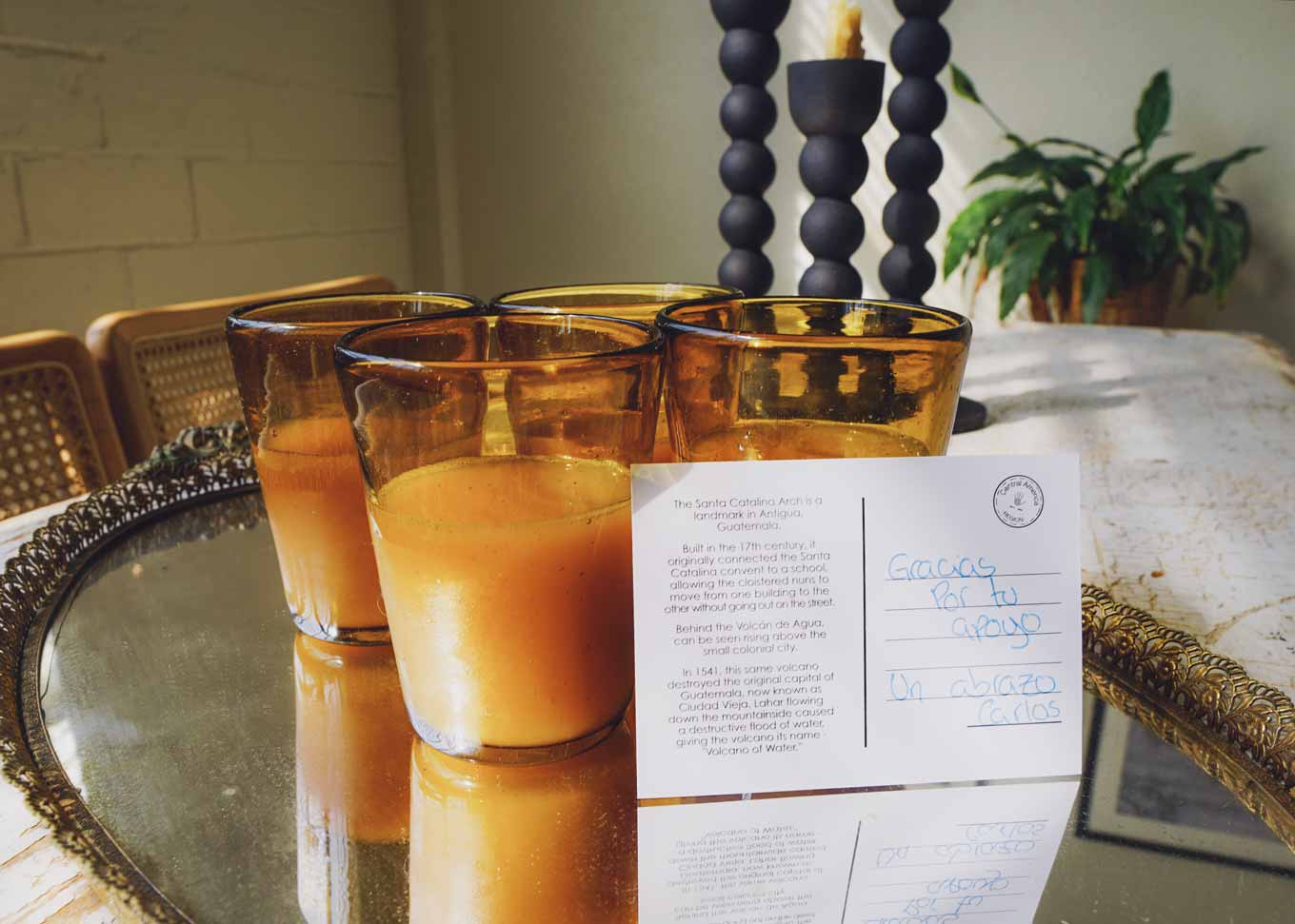 Four hand-blown recycled drinking glasses in amber on a tray at a table, with a note from the maker written in Spanish