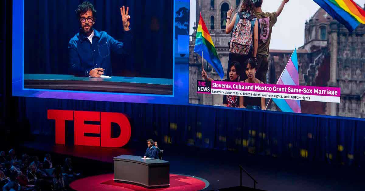 A white man with brown curly hair and glasses holds up three fingers as he speaks on the TED stage. Behind him are two large monitors.