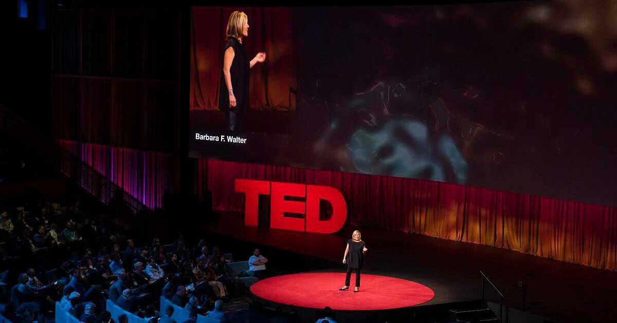 A white woman wearing all black stands on the TED stage in front of an attentive audience.