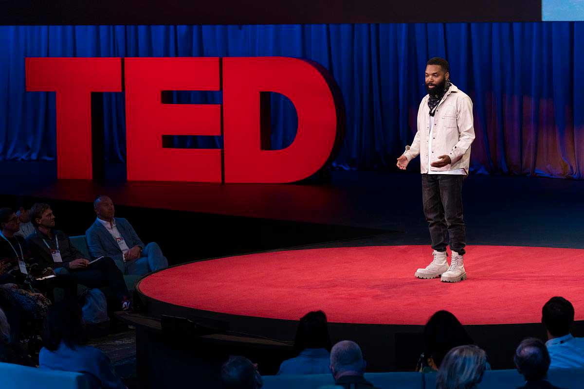 A Black man in a white jacket and black pants speaks on the TED stage.