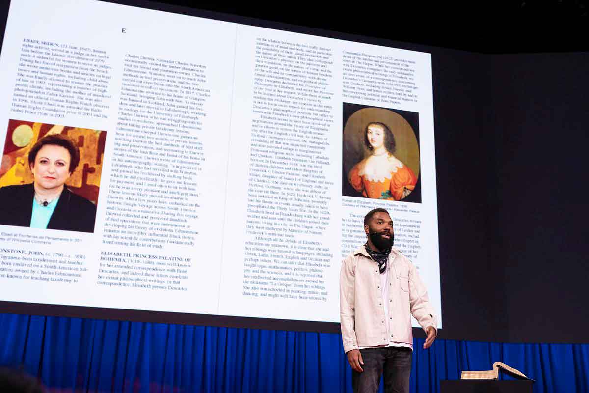 A Black man in a white jacket and black pants speaks on stage, a screen displaying pages of an encyclopedia behind him. 
