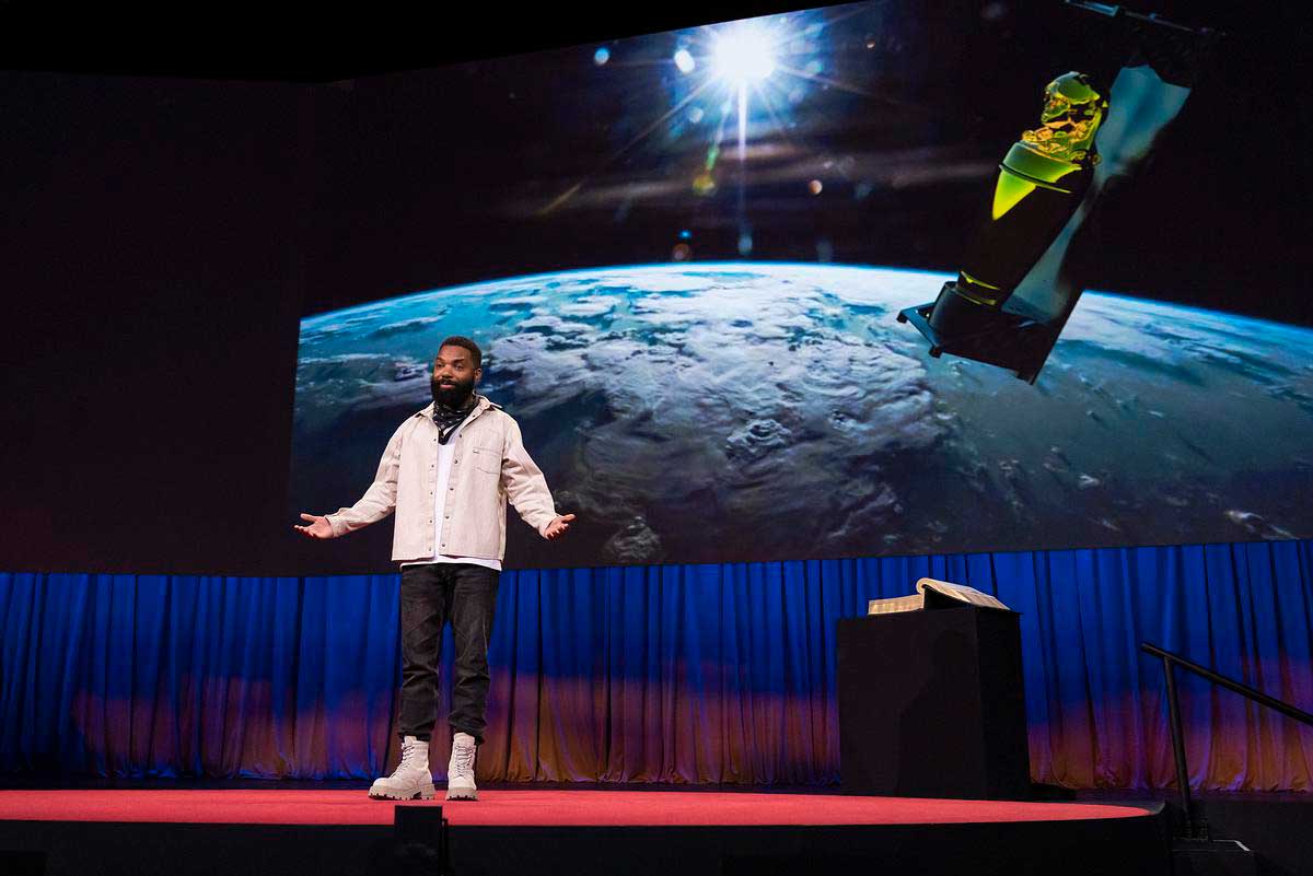 A Black man in a white jacket and black pants speaks on stage, a photo of a satellite displayed on a screen behind him. 