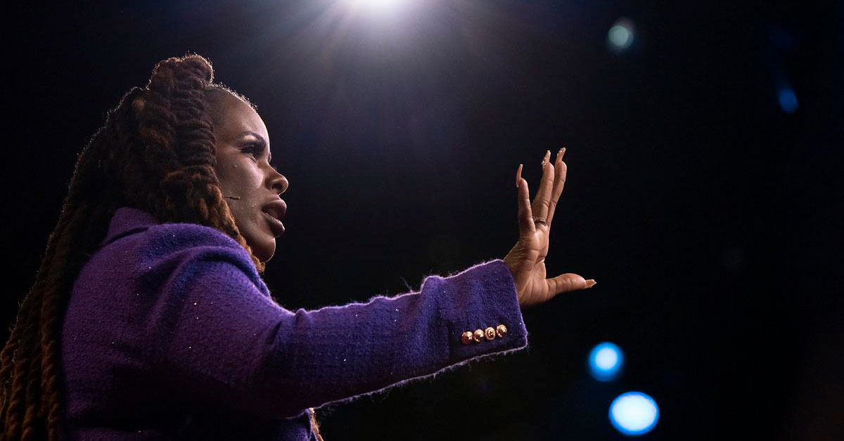 A Black woman with braids and a purple blazer extends her arm out to a crowd as she speaks on stage.