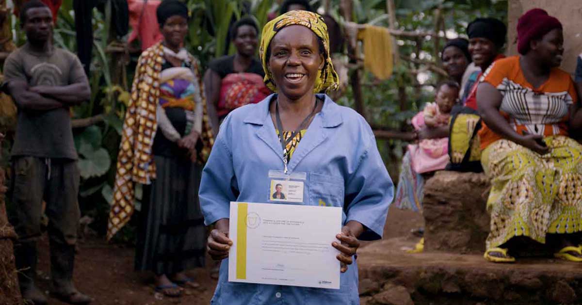 An African woman wearing a blue shirt and yellow headpiece holds up a certificate, smiling. In the background, other community members mill about. 