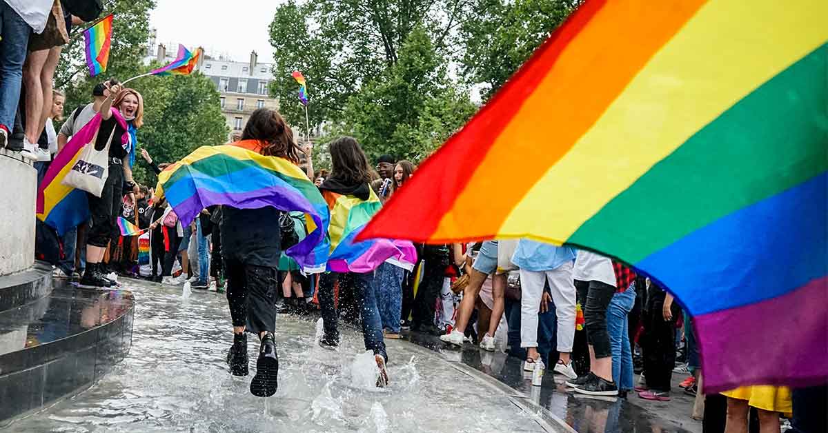 Two people run in a fountain wearing rainbow flags as capes while people around them wave rainbow flags