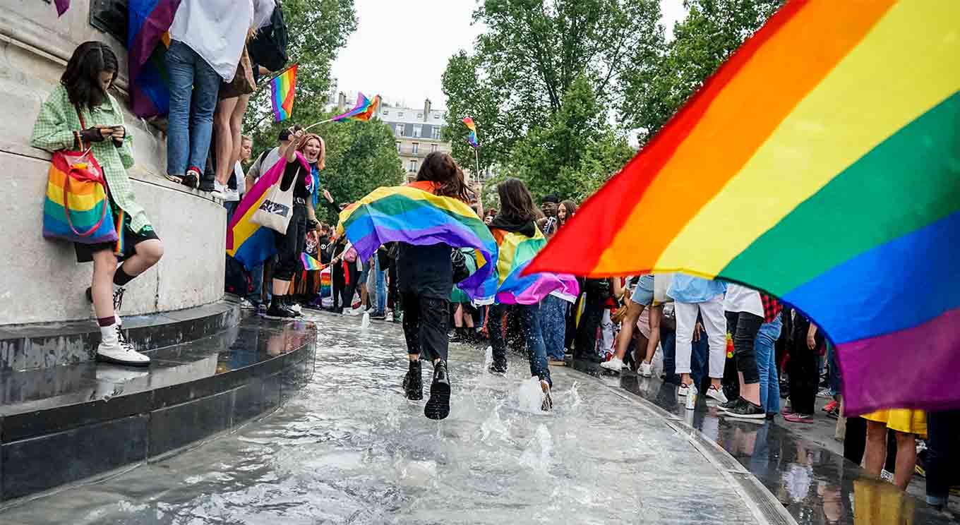Two people run in a fountain wearing rainbow flags as capes while people around them wave rainbow flags