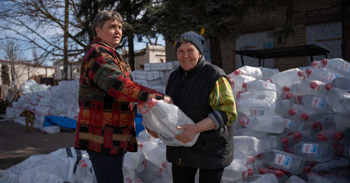 A Ukrainian is handed a gallon of drinking water. Palettes of water are in the background.