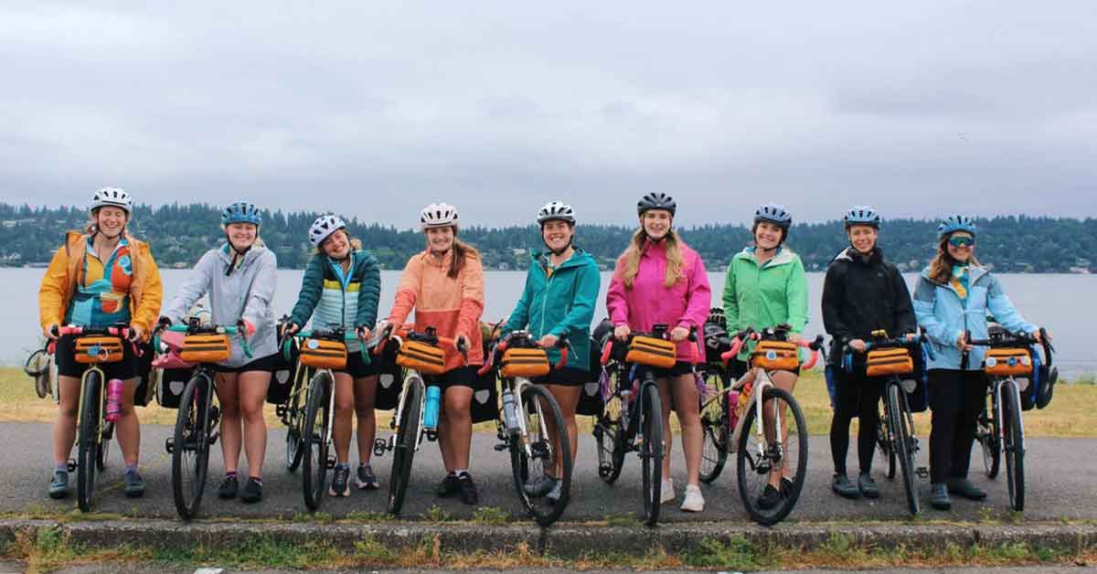 Nine young women stand next to their bicycles, cloudy skies and a body of water behind them.
