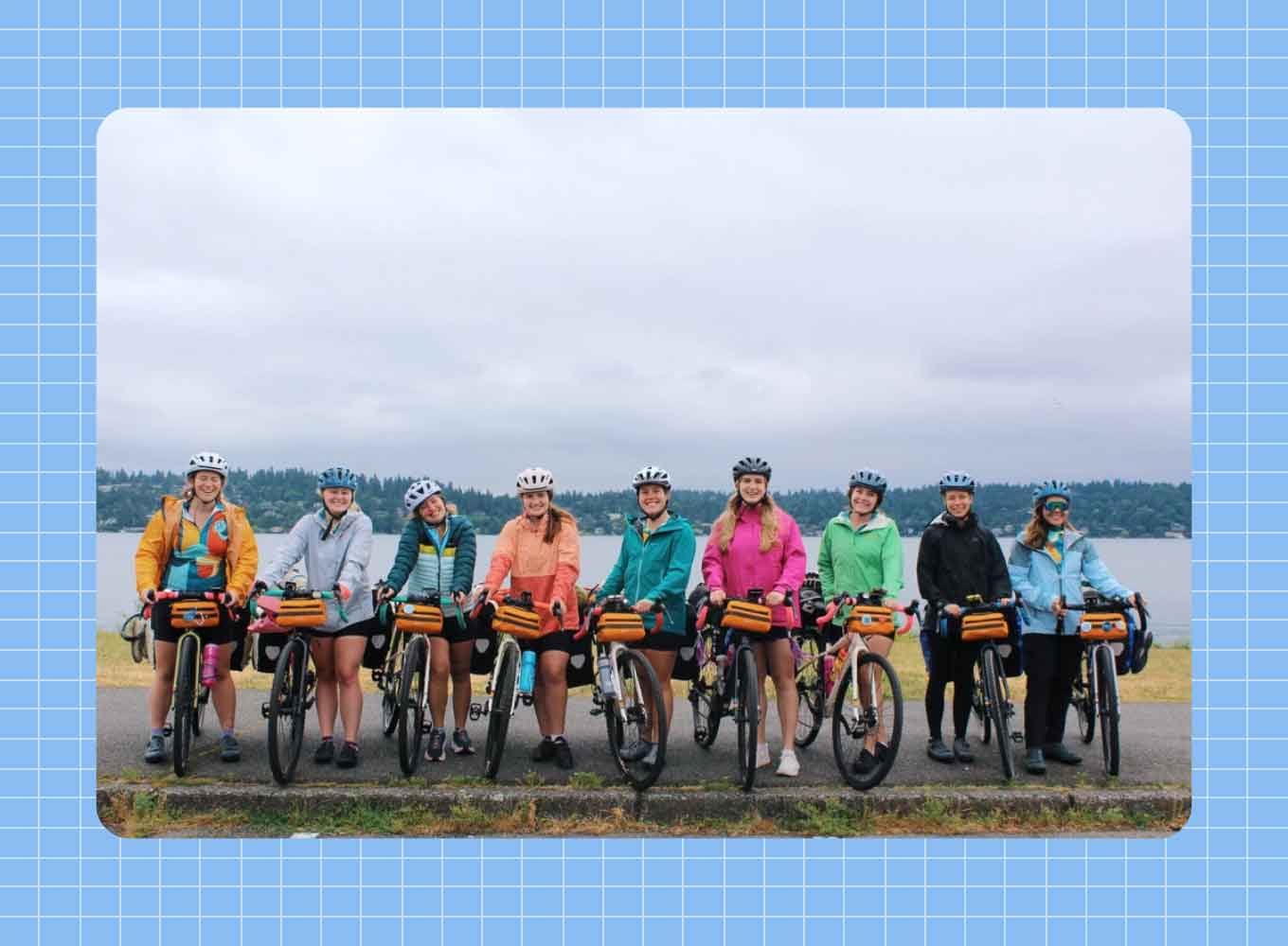 Nine young women stand next to their bicycles, cloudy skies and a body of water behind them.