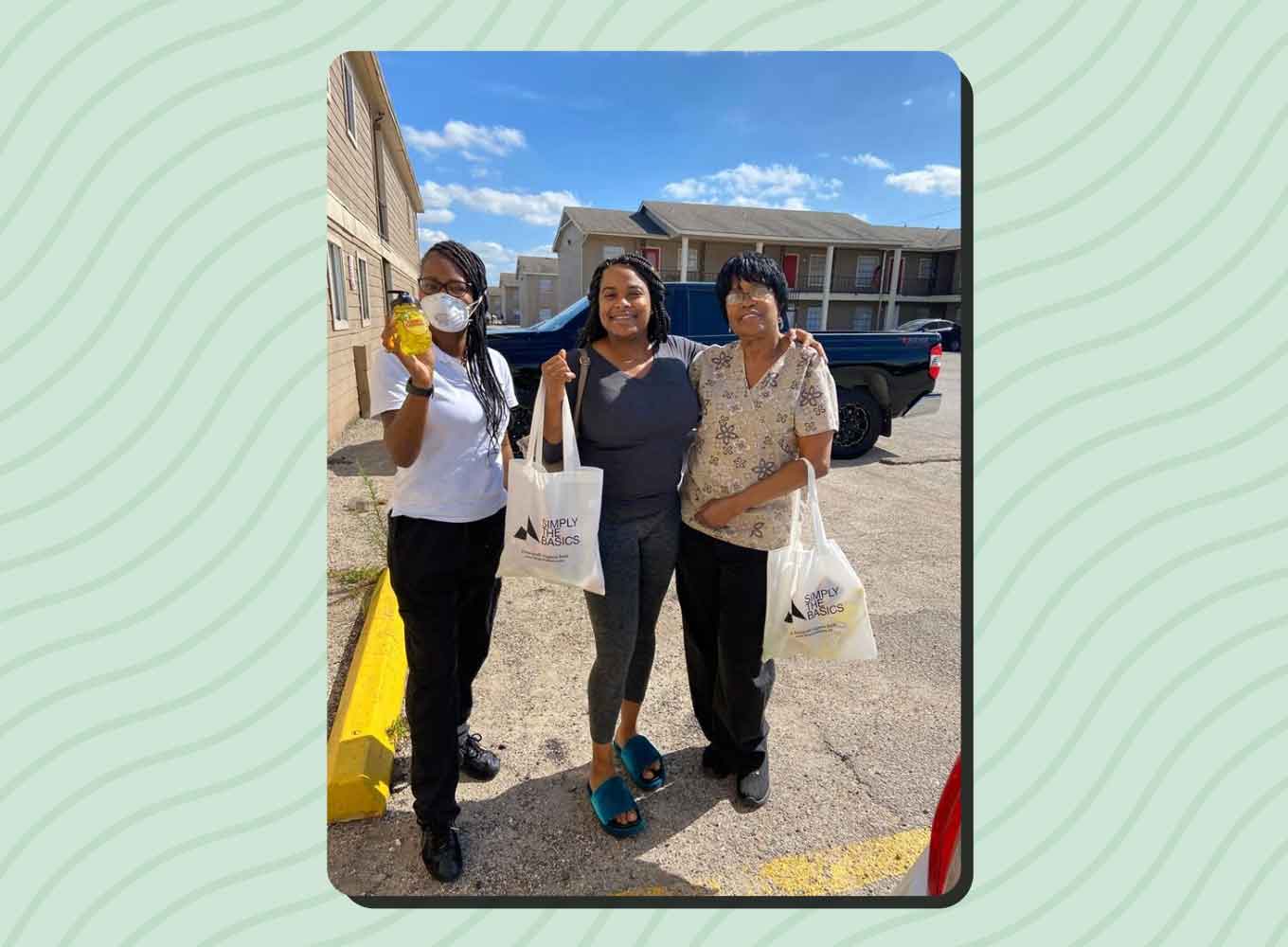 Three Black women stand in a parking lot with Simply the Basics tote bags. One holds up a bottle of soap. 