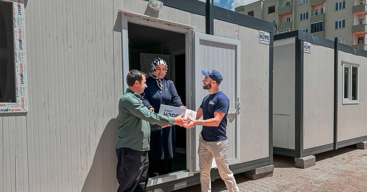 Two men hand a woman a box in the doorway of a housing container
