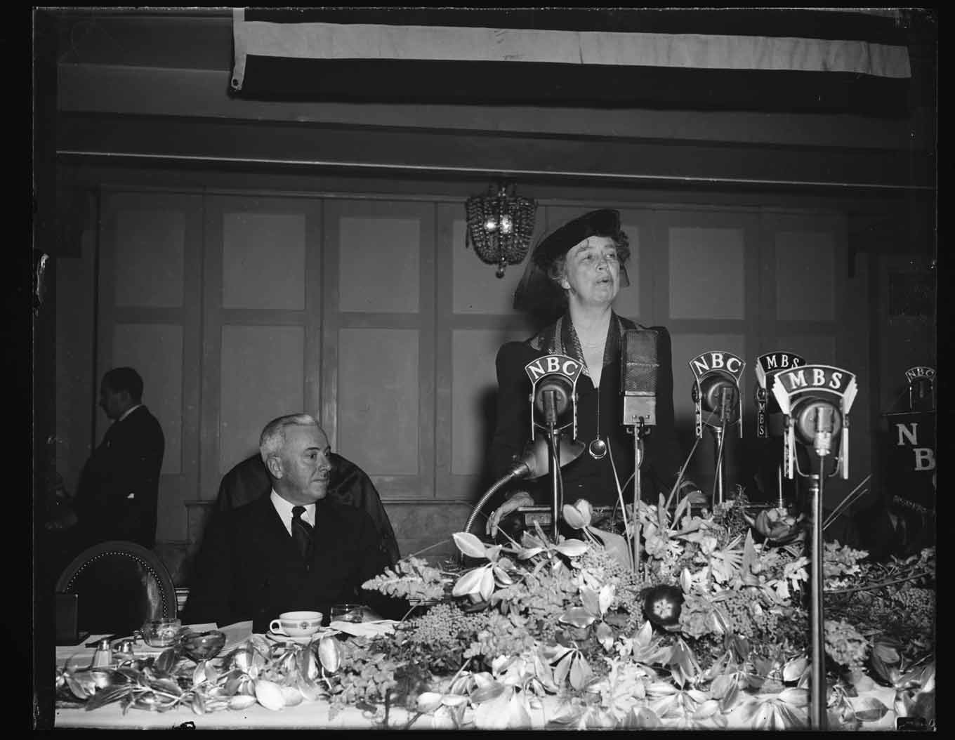 Eleanor Roosevelt talking in a press conference while standing up with a seated Franklin D. Roosevelt at her side