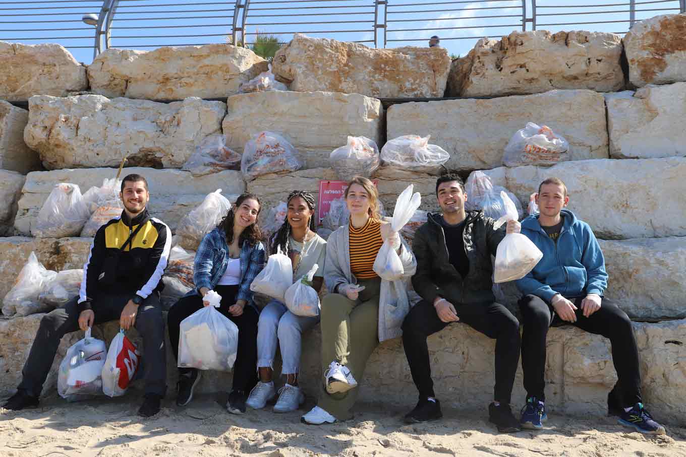 A group of 6 people sit on large rocks, holding up bags of trash.