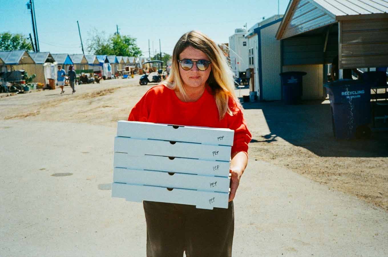 A woman with blonde hair wears a red crewneck sweatshirt and black sunglasses while holding five boxes of pizza