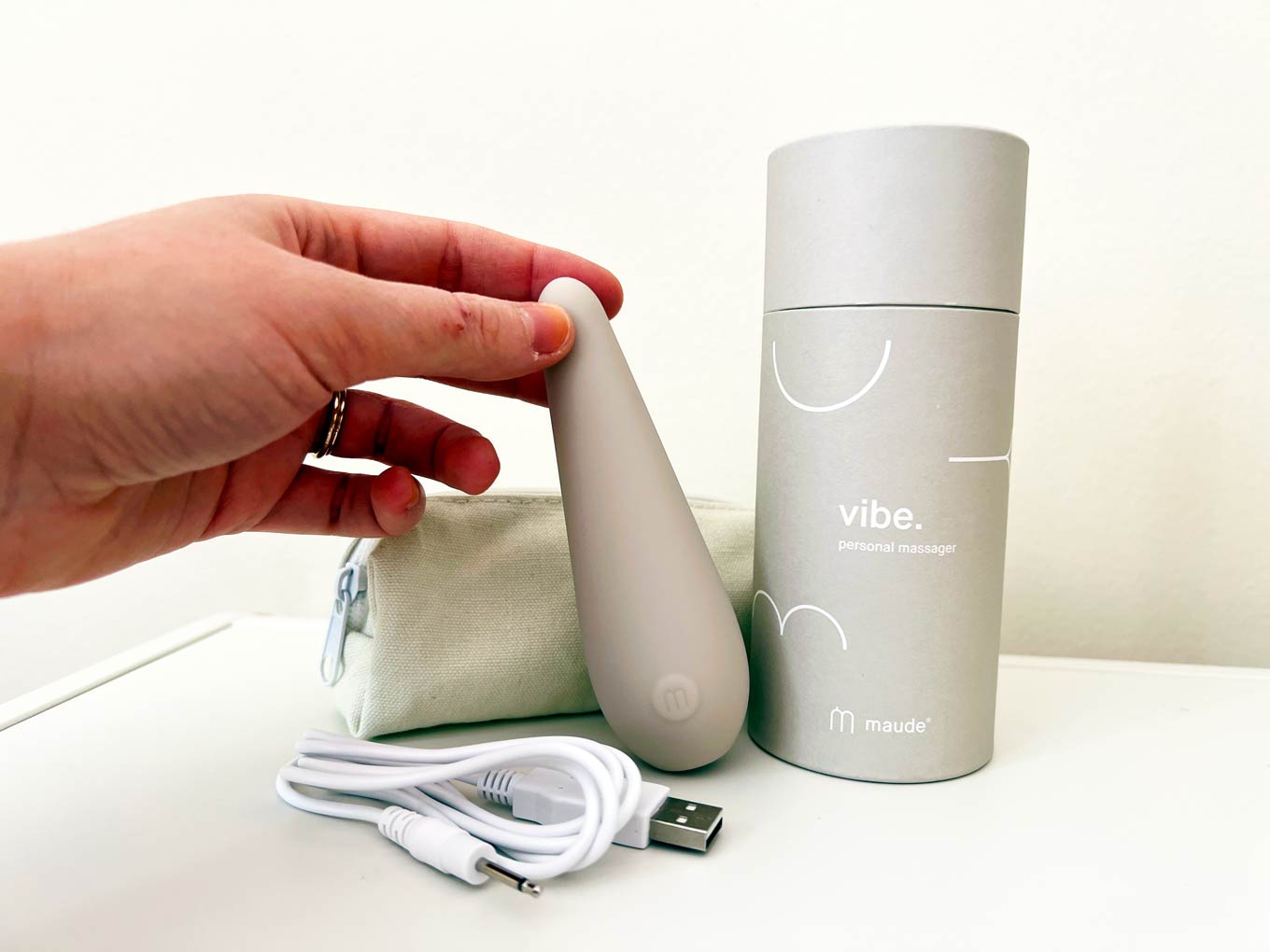 A hand holds up the Vibe from Maude, next to its packaging and carrying case